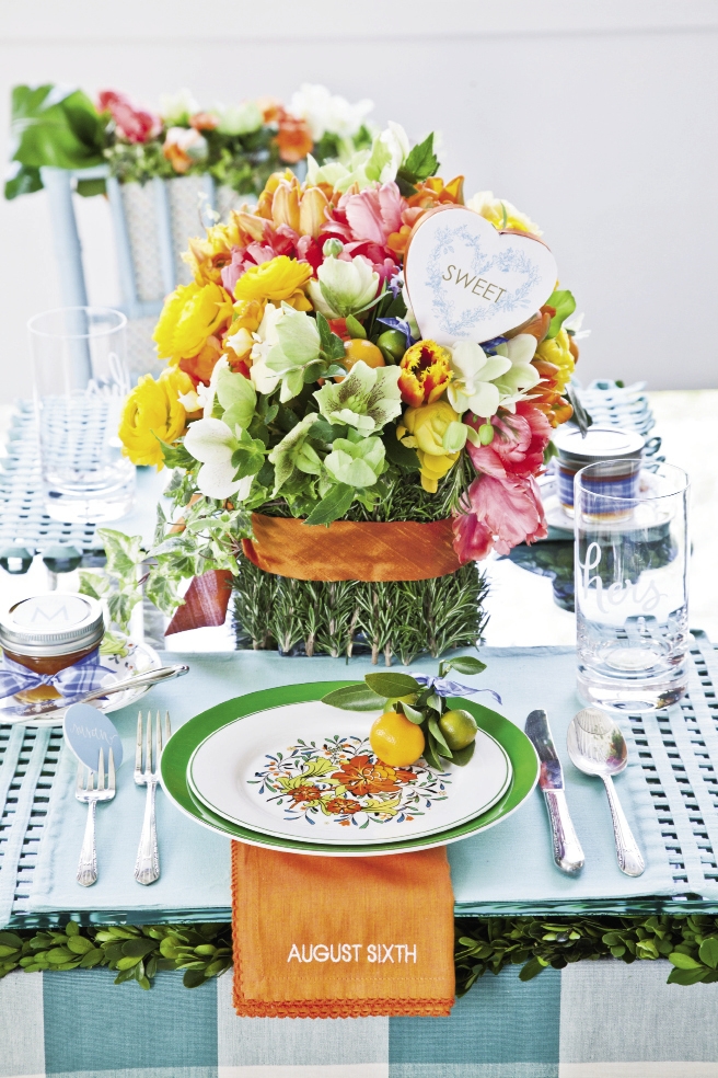 POLISHED PLACESETTING: Anne placed boxwood mats from Jamali Garden on the table, then topped those with glass from Charleston Architectural Glass Studio. She then fashioned florals from coral begonia, bird’s nest fern, green hellebores, variegated ivy, chartreuse phalaenopsis orchids, orange and gold ranunculus, rosemary, and tulips. Simple bands of Thai silk ribbon accented the arrangement and bouquet.