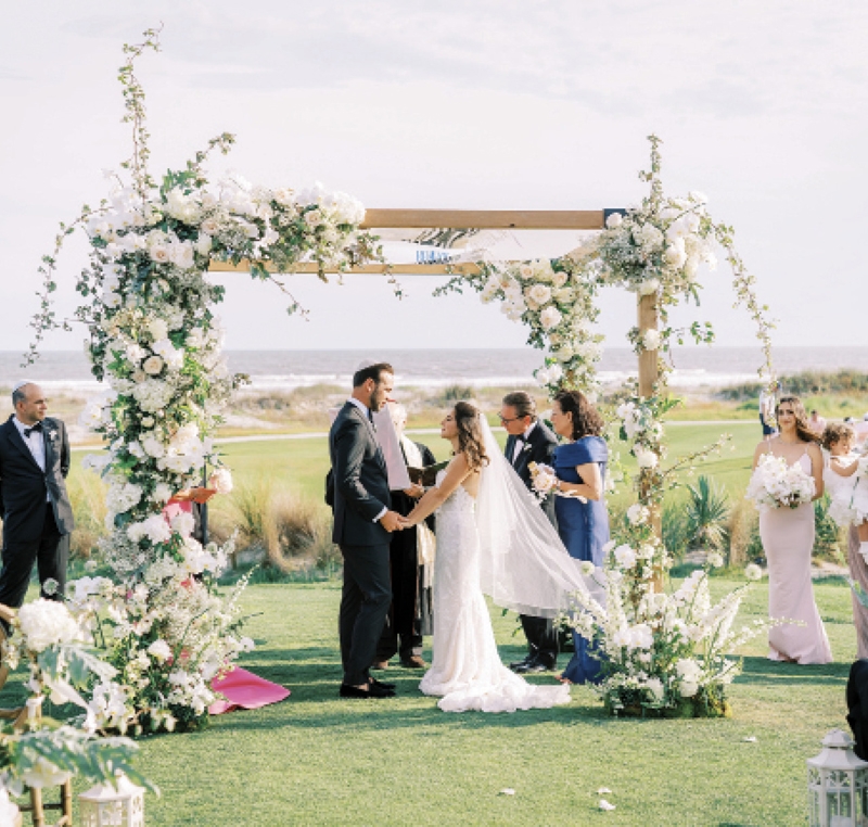 The couple married beneath a floral-festooned chuppah, its crisp white blooms reflected in Sarah’s lace Netta BenShabu dress.